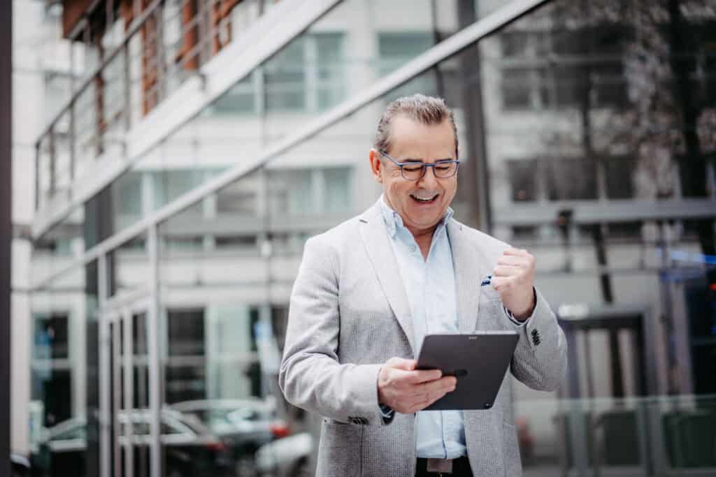 corma- A man in a light gray blazer looks at a tablet, smiling and raising one fist in excitement. He stands outside a glass building.